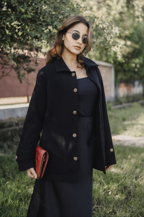 Woman in a black coat and sunglasses standing outdoors with trees in the background